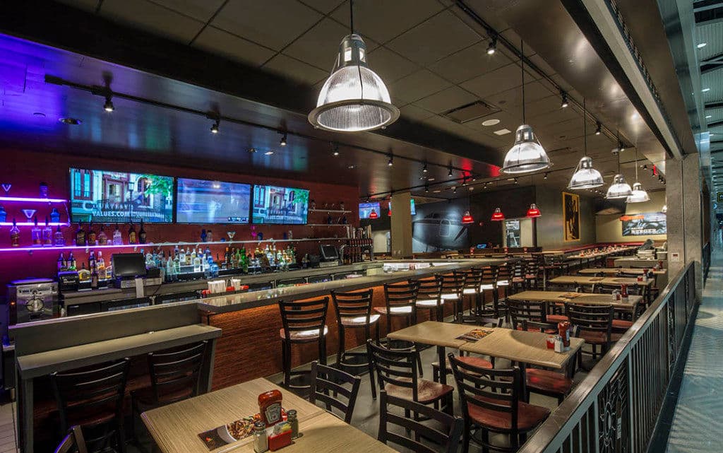 Modern restaurant bar with illuminated shelves, seating, and screens displaying menus.