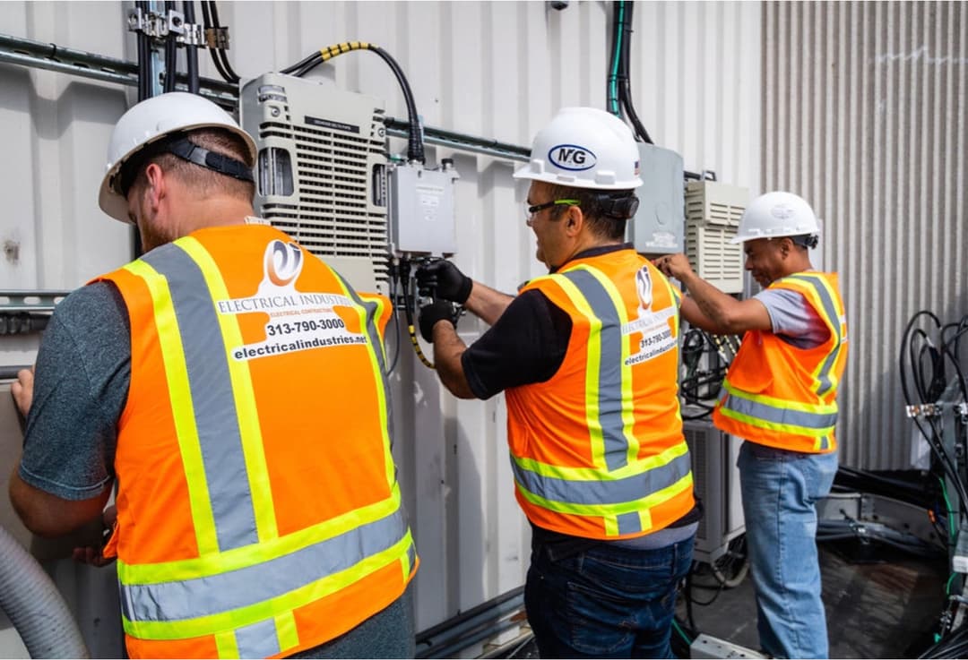 Three workers in safety vests and hard hats repairing electrical equipment outdoors.