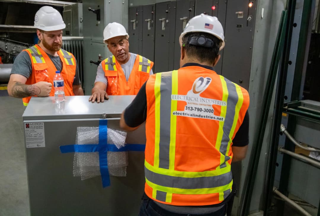 Workers in hard hats and safety vests collaborating on an electrical panel installation.